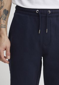 Navy sweatpants with an elastic waistband, drawstring ties, side pockets, and a soft texture. Features visible stitching and metallic accents.