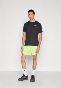 Μαύρο κοντομάνικο μπλουζάκι Nike, νέον πράσινα σορτς, μαύρα αθλητικά παπούτσια, λευκές κάλτσες με το λογότυπο Nike, στέκεται μπροστά σε απλό φόντο.