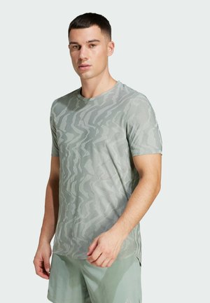 Giovane uomo che indossa una camicia a maniche corte grigio con un motivo e pantaloni corti verde chiaro, in piedi su uno sfondo chiaro e neutro.