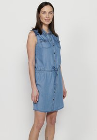 Abito senza maniche in denim blu chiaro con chiusura a bottoni sul davanti, colletto, due tasche sul petto e vita con cordoncino. Decorato con dettagli a spalla a fantasia.