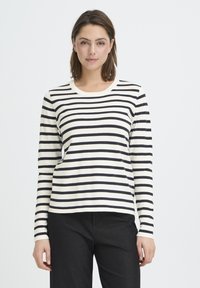 Haut à manches longues crème à rayures horizontales noires, tissu en coton lisse, encolure ronde et silhouette ajustée. Associé à un pantalon noir.