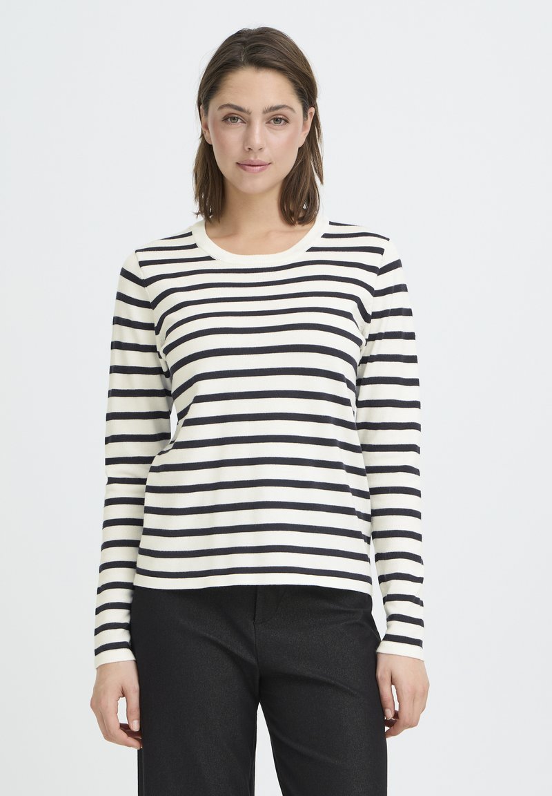 Haut à manches longues crème à rayures horizontales noires, tissu en coton lisse, encolure ronde et silhouette ajustée. Associé à un pantalon noir.