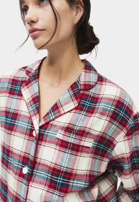 Pigiama a scacchi in morbido cotone con tonalità di rosso, blu e grigio. Presenta una chiusura con bottoni, colletto a punta e taschino sul petto.