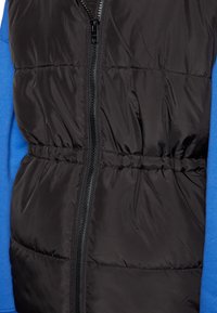 Czarna kamizelka puchowa z zapięciem na zamek i elastycznym paskiem w talii, nałożona na niebieską koszulę z długim rękawem. Gładka tekstura, bez widocznych wzorów.