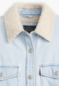 Lyseblå denimjakke med en fluffy beige krage, to frontlommer, metallknapper og subtile sømdetaljer.