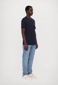 Tamsiai mėlyna trumpa rankovė T-shirt su apvaliu iškirpimu, dėvima su šviesiai mėlynomis džinsinėmis kelnėmis ir baltais sportbačiais. Paprastas dizainas, be raštų.