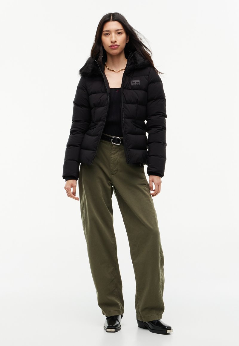 Femme portant une doudoune noire avec un col en fourrure, un haut noir, un pantalon large vert olive, une ceinture noire et des bottes noires à talons, debout sur un fond blanc.
