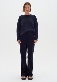 Naine seisab, seljas tumm sinine soe kampsun, laiad tumehalli püksid ja mustad sandaalid, taustal on ühtlane valge taust.