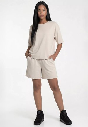 Kobieta o długich czarnych włosach, ubrana w beżowy top z krótkim rękawem i szorty oraz czarne wysokie trampki, stojąca z rękami w kieszeniach.