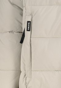 Beige gesteppter Stoff mit genähten Nähten und einem kleinen schwarzen rechteckigen Etikett mit der Aufschrift "SUBLEVEL" neben einem schwarzen Schlaufen-Zugtab.