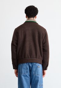 Brun tekstureret jakke med en ribbet krave med grønne og hvide striber, parret med lyseblå jeans. Jakken har en afslappet pasform.