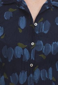 Námornícky kvetinový krátky rukáv s modrými tulipánovými vzormi a zelenými listami. Má golier s gombíkmi a biele gombíky na textúrovanej tkanine.