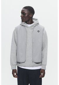 Grijze mouwloze vest over een grijze hoodie. Het vest heeft een rits aan de voorkant en een logo op de borst. Soepele stof, lichtgewicht design, relaxte pasvorm.
