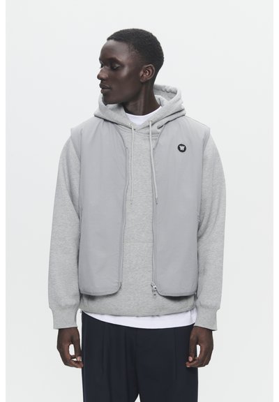 Gilet sans manches gris par-dessus un hoodie gris. Le gilet a une fermeture éclair à l'avant et un logo sur la poitrine. Tissu lisse, design léger, coupe décontractée.