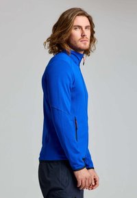 Uomo con capelli lunghi fino alle spalle che indossa una giacca blu con zip e pantaloni scuri, in piedi di profilo su uno sfondo semplice.