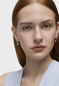 Orecchini a cerchio in argento con un piccolo pendente pendente, con una finitura liscia. Il soggetto ha capelli lisci di un colore rosso-bruno e una pelle chiara.