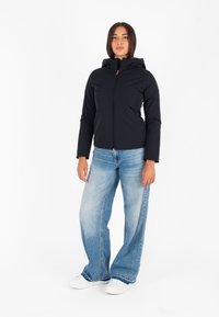 Giacca nera con cappuccio e zip frontale, dotata di tasche laterali e pannelli testurizzati. Abbinata a jeans a gamba larga blu chiaro e sneakers bianche.
