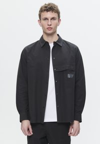 Black button-up shirt with a chest pocket and contrasting stitching. Features a logo detail and long sleeves. Worn over a white t-shirt.