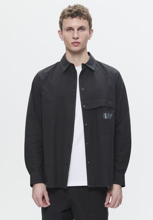 Black button-up shirt with a chest pocket and contrasting stitching. Features a logo detail and long sleeves. Worn over a white t-shirt.