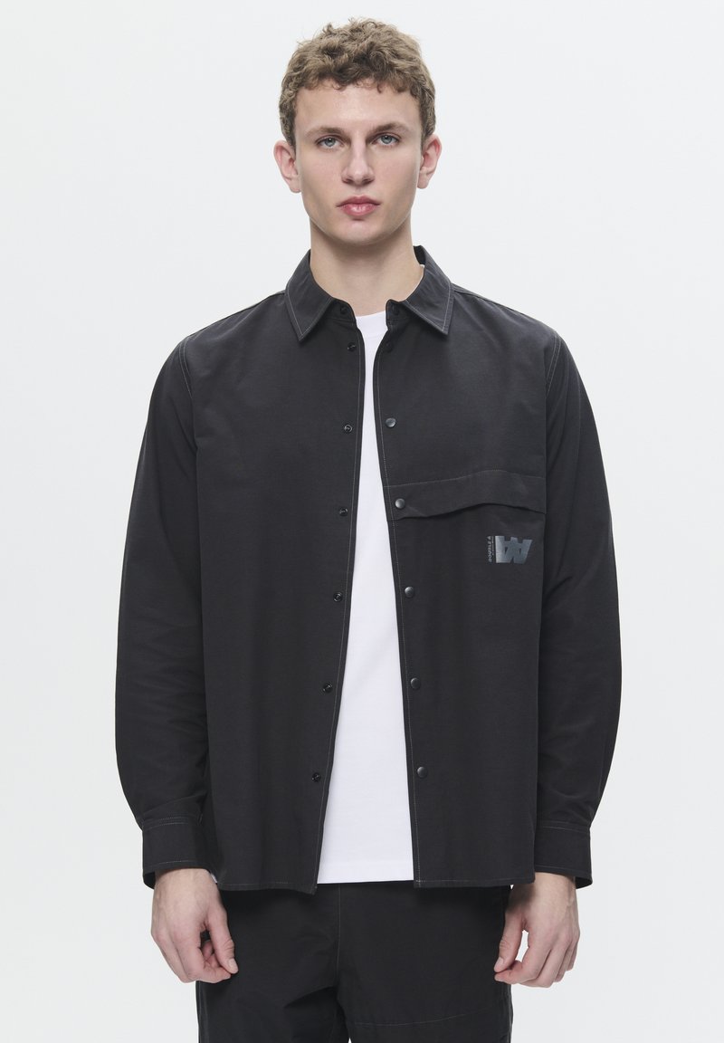 Black button-up shirt with a chest pocket and contrasting stitching. Features a logo detail and long sleeves. Worn over a white t-shirt.