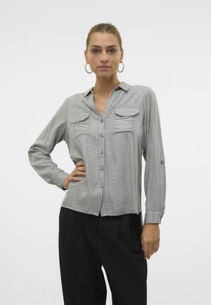 Mujer con cabello castaño claro, con una camisa gris abotonada con rayas verticales y pantalones negros, de pie con una mano en la cadera.