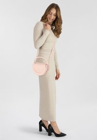 Bolso cruzado de cuero rosa con un diseño curvado, correa ajustable y sutil logotipo en relieve, llevado con un vestido beige y tacones negros.