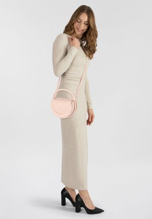 Borsa crossbody rosa in pelle con design curvo, tracolla regolabile e logo in rilievo discreto, indossata con un vestito beige e tacchi neri.