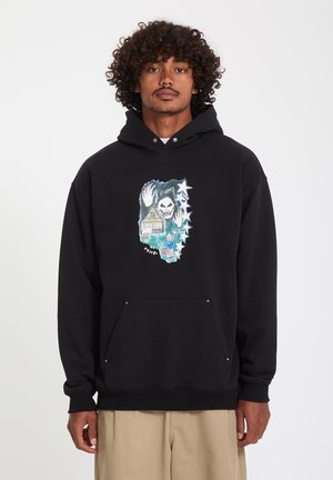Homme aux cheveux bouclés portant un sweat à capuche noir avec un graphique représentant un crâne, des étoiles, une maison et une personne saluant, avec le texte "Volcom" à l'avant.