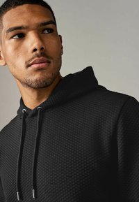 Zwarte hoodie van textuur met een diamanten patroon, voorzien van een trekkoord capuchon en een slank ontwerp, gedeeltelijk weergegeven op de bovenste torso van een model.