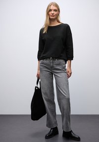 Top lungo a maniche lunghe nero con texture abbinato a jeans grigi a gamba dritta a vita alta. Il modello tiene una borsa a tracolla in suede nera e indossa stivaletti neri.