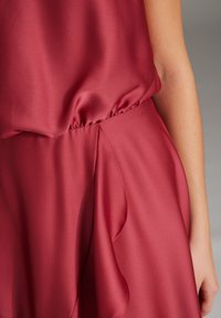 Robe en satin rouge avec une taille froncée, un drapé doux et des détails de jupe fluide. Le tissu a une texture lisse et un éclat subtil.