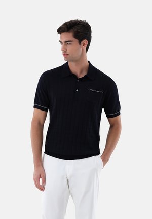 Marinefarbener Polo-Shirt mit kurzen Ärmeln, strukturiertem Muster, dekorativer Stickerei am Kragen und einer kleinen Brusttasche. Getragen mit weißen Hosen.
