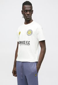 Bílé tričko s krátkými rukávy s kulatým květinovým motivem, žlutými akcenty a černým textem. Kombinováno se šedými tepláky.