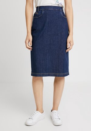 Mujer con una falda de mezclilla azul oscuro hasta la rodilla con bolsillos, combinada con zapatillas blancas y una blusa blanca con estampado floral.