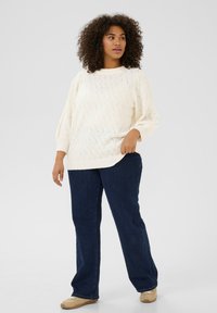 Pull en maille crème avec motif texturé, associé à un jean large bleu foncé et des chaussures beiges. Tissu doux avec une coupe décontractée.