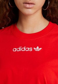 Röd bomullströja med vit broderad "adidas" logotyp och tre ränder logotyp på framsidan, med en klassisk rund halsringning.