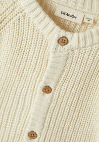 Kreminės spalvos minkštai megztas kardiganas su raukšlėta apykakle, prisiūtomis medinėmis sagomis ir tekstūruotu audiniu. Etiketė matoma viduje.