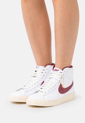 Sneakers bianchi alto con accenti bordeaux, caratterizzati da un materiale testurizzato, lacci classici e un piccolo dettaglio decorativo a catena.