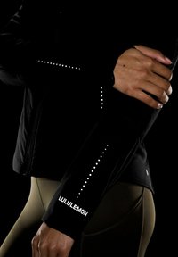 Crna atletska jakna s reflektirajućim točkama na rukavima, s bijelim Lululemon logom; tkanina izgleda glatko i prilagođeno.