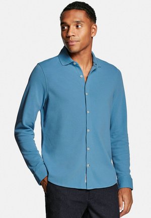 Homme portant une chemise bleue à manches longues boutonnée avec col, debout les mains dans les poches, regardant sur le côté contre un fond uni.
