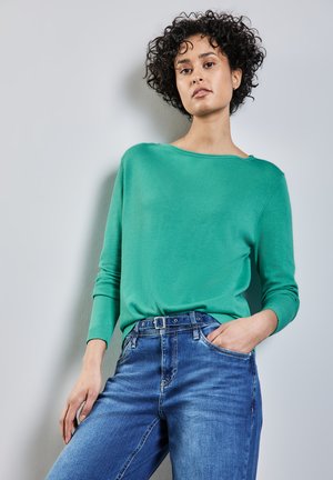 Grön tröja med rundad halsringning och långa ärmar, tillsammans med blå jeans som har ett bälte och framfickor. Slät textur.