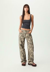 Mörkgrå tajt linne som matchas med lösa jeans i kamoflage-mönster i olivgrön och tan. Modellen bär bruna platåsandaler och har händerna i fickorna.