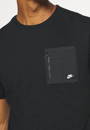 Black T-shirt with short sleeves, featuring a textured pocket with a zipper and a small Nike logo on the front. Smooth cotton material.