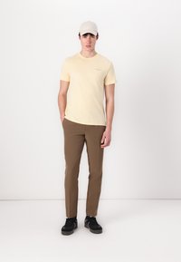 T-shirt jaune clair avec un petit logo textuel, pantalon slim marron, et baskets noires. Modèle portant une casquette gris clair, se tenant sur un fond blanc.