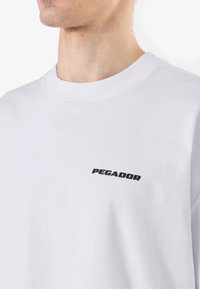 Läheltä kuva henkilöstä, joka käyttää valkoista crewneck-paitaa, johon on printattu mustalla "PEGADOR" vasempaan rintaan.