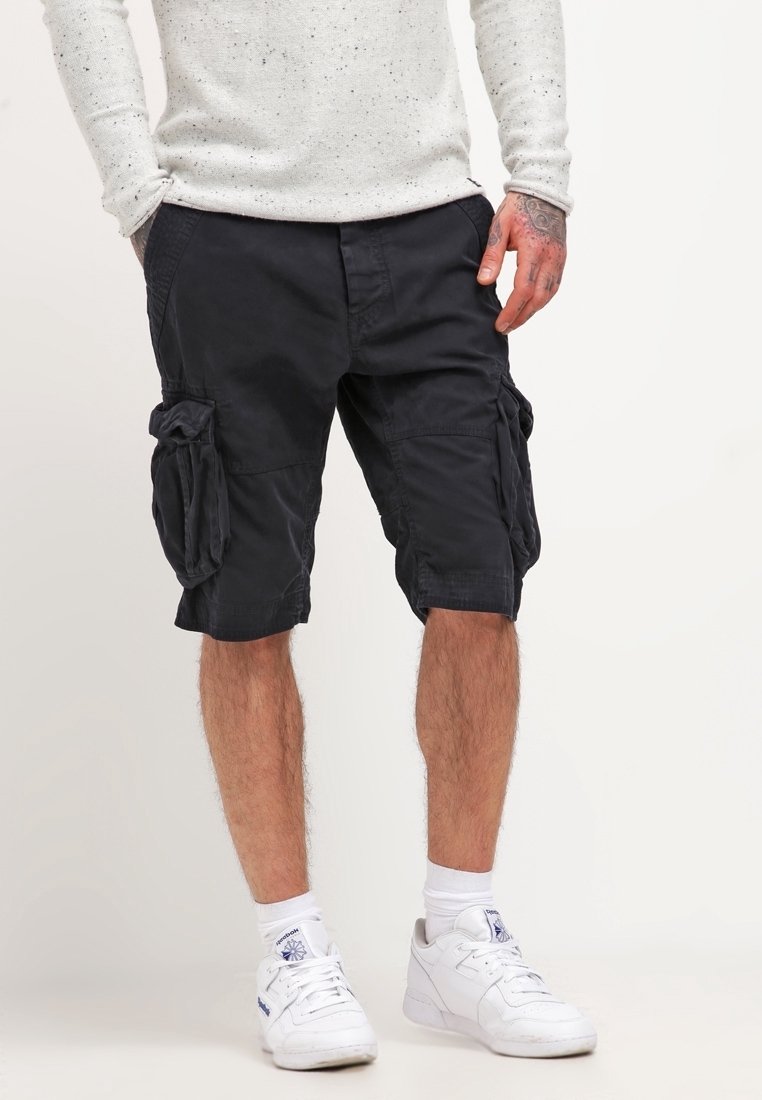Homme portant un short cargo noir, des chaussettes blanches et des baskets blanches, avec une main dans la poche et la main tatouée visible.