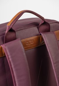 Zaino di tessuto viola con dettagli in pelle marrone, dotato di maniglia superiore e spallacci imbottiti. Etichetta visibile sul retro.