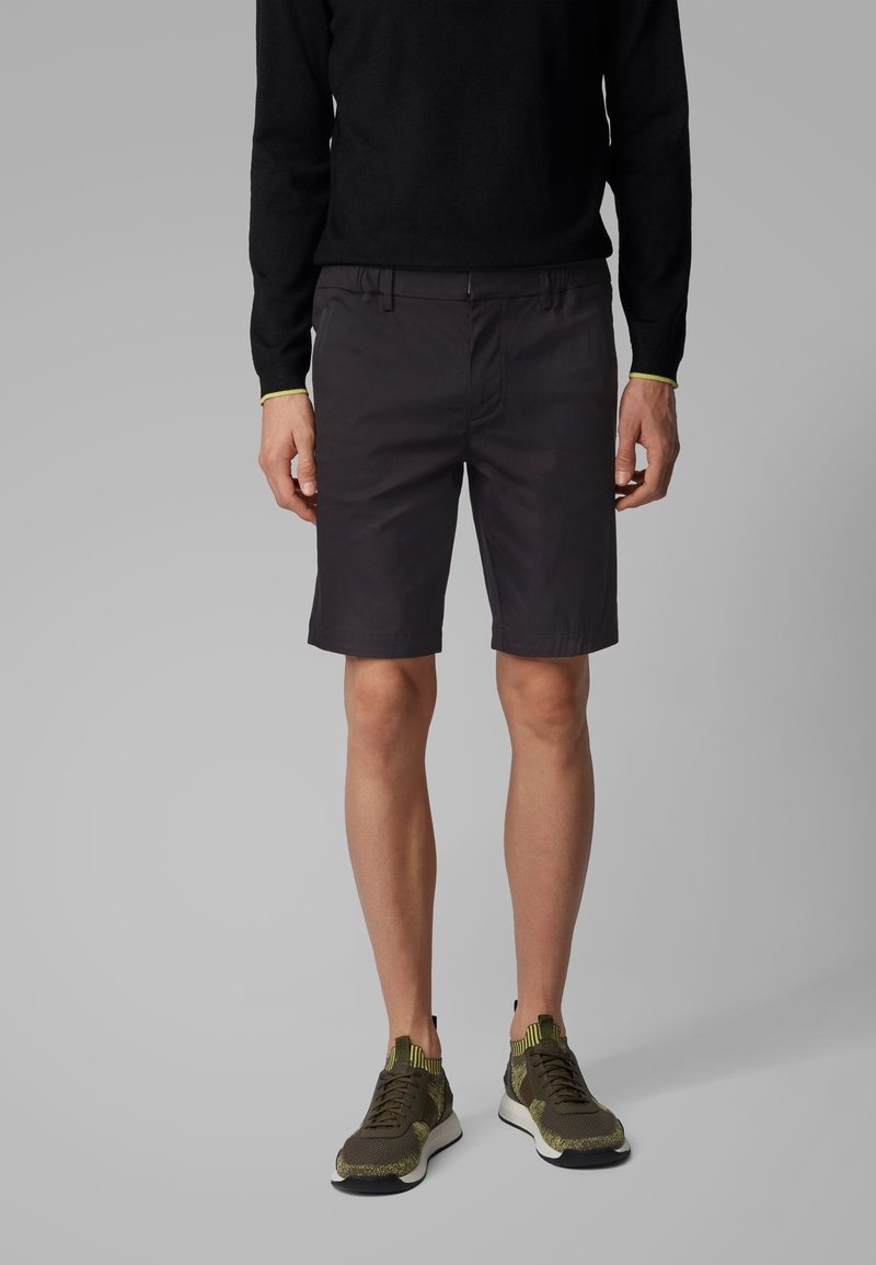 Pull noir à manches longues, short foncé et baskets vertes. Le short présente un design à devant plat, des poches latérales et une coupe ajustée.