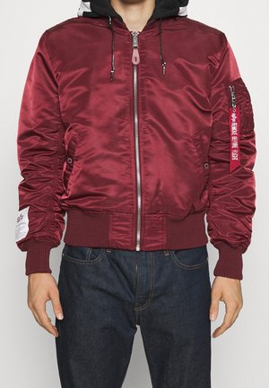 Kastanienbraune Bomberjacke mit glatter, glänzender Oberfläche, gerippten Bündchen und Saum, Reißverschluss, Seitentaschen und einem roten Etikett am Ärmel.