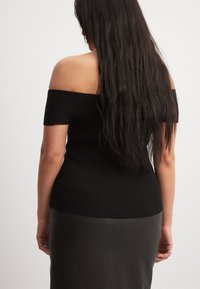 Top nero a spalla scoperte con un design aderente, abbinato a una lunga gonna scura e dalle texture ricca. Capelli che cadono dritti lungo la schiena.